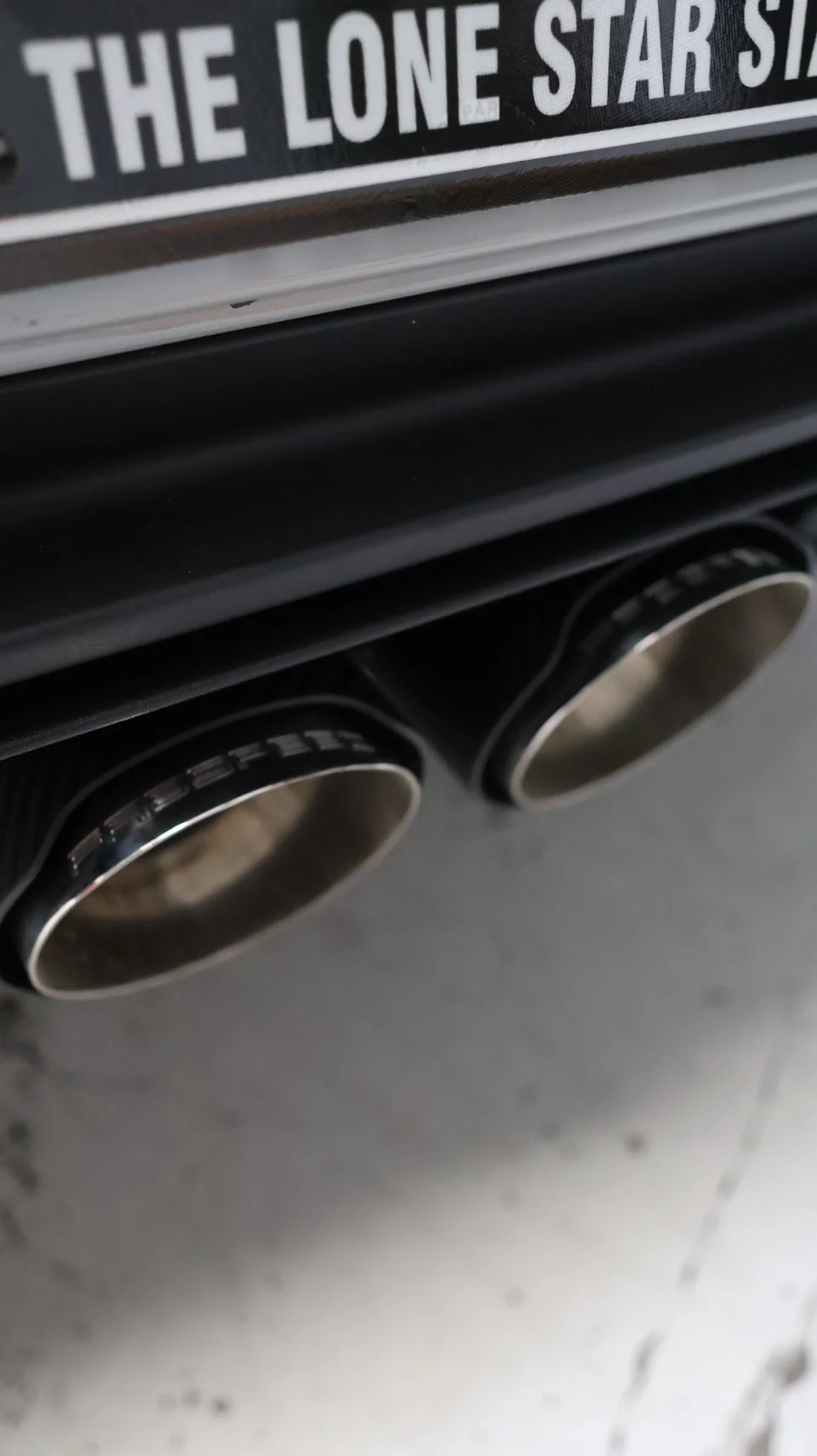 Close-up of a car's dual exhaust tips with a carbon fiber-like trim and a partial view of a license plate.