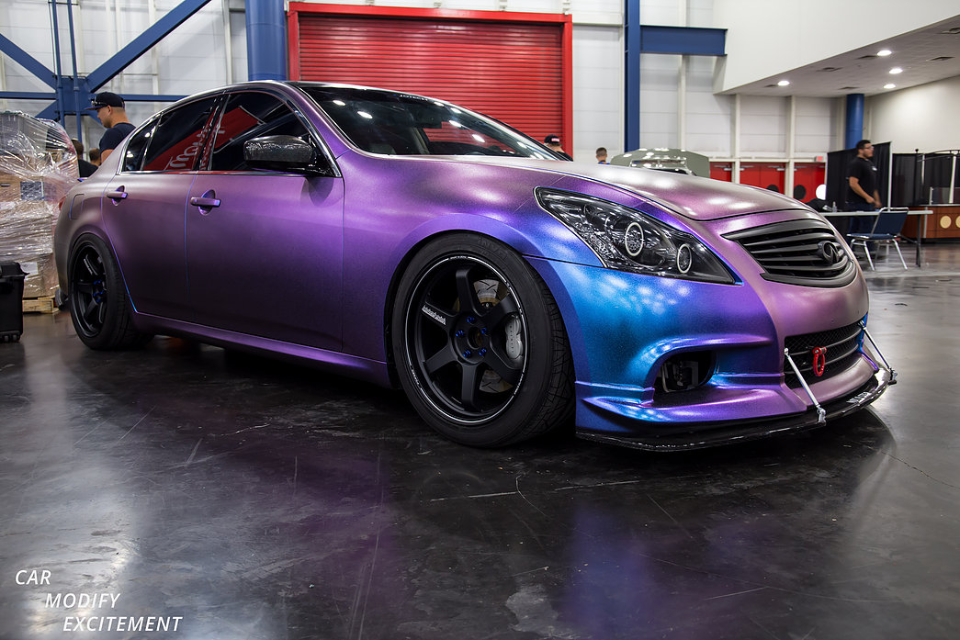 Custom Infiniti G37 sedan with a vibrant chameleon wrap, aftermarket wheels, and a front splitter displayed at a car show.