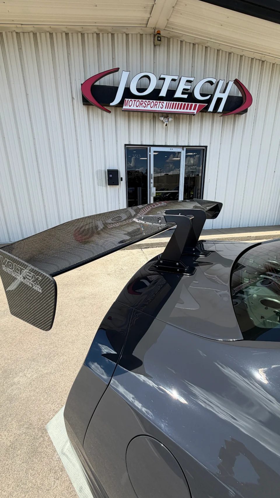 A dark grey sports car with a large Voltex carbon fiber rear wing parked outside Jotech Motorsports shop.