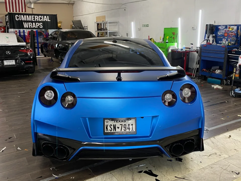 Rear view of a matte blue wrapped Nissan GT-R R35 with a black spoiler and quad exhaust tips in a car shop.