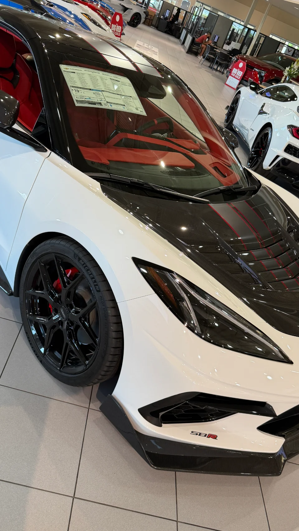 A white Chevrolet Corvette C8 with black carbon fiber accents, black wheels, and a red interior on display in a showroom.