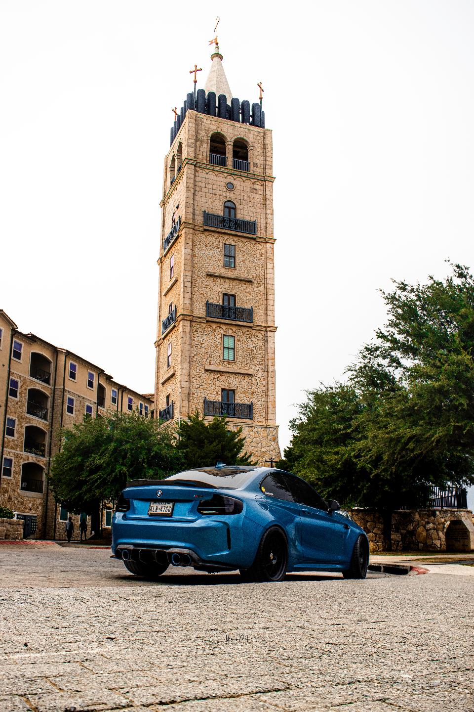 A blue BMW M2 with aftermarket wheels and custom exhaust parked in front of a tall stone tower and building.