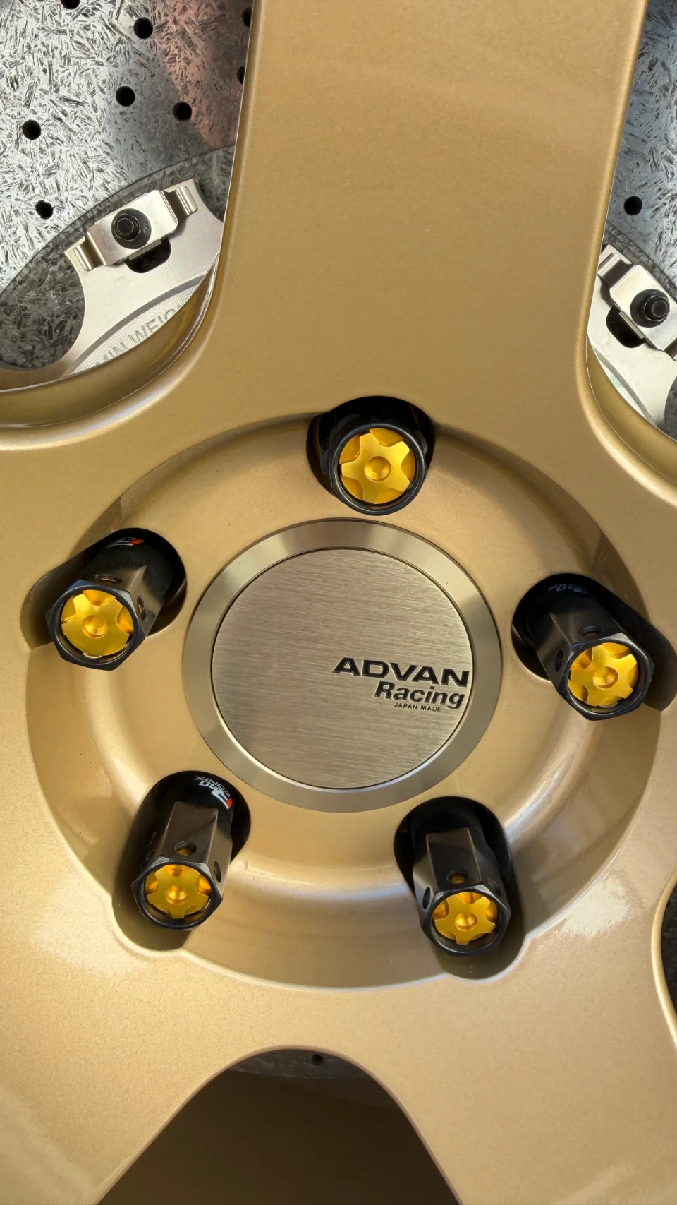A close-up of a gold Advan Racing wheel with black and gold lug nuts and a perforated brake rotor.
