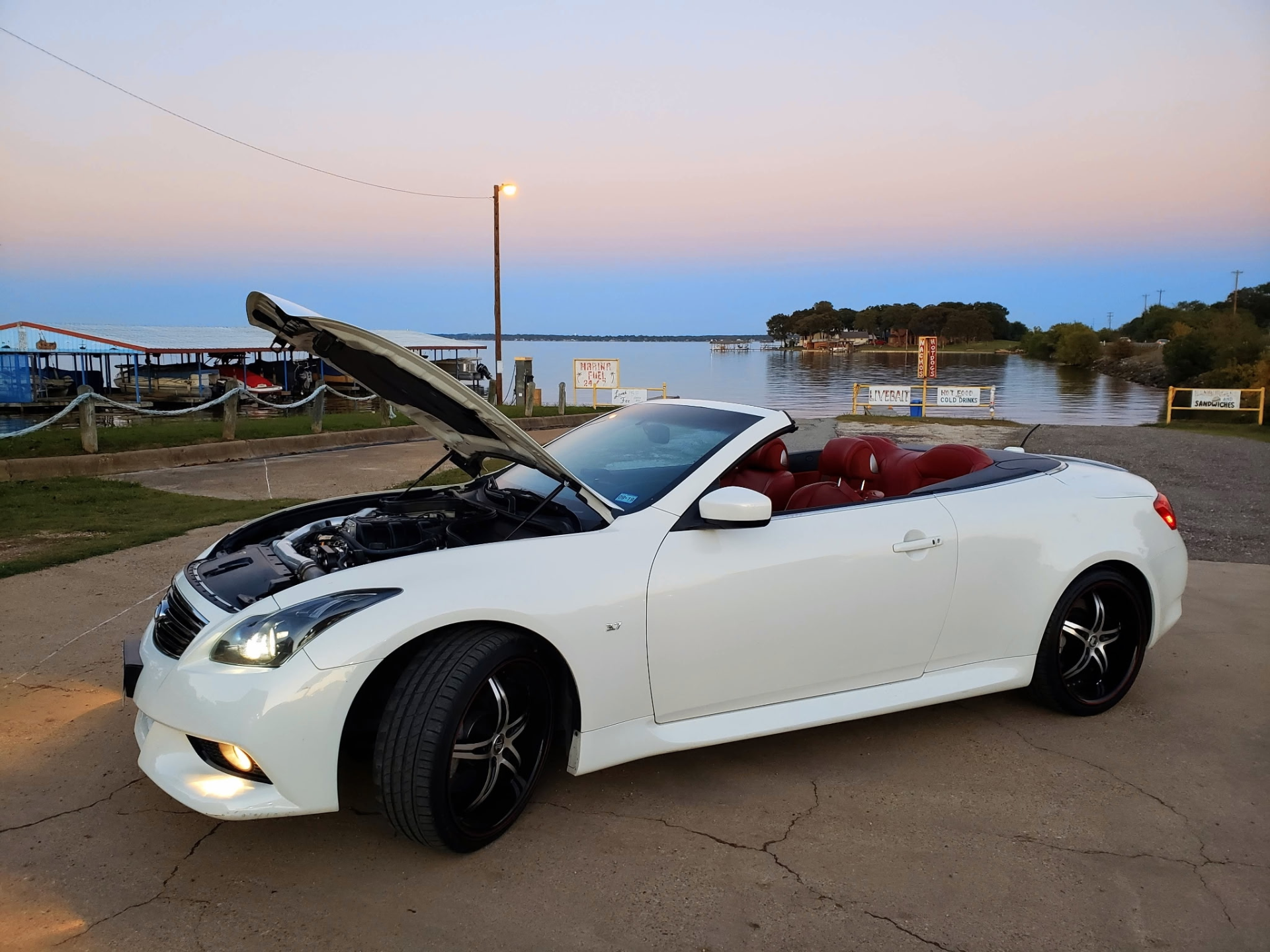 White Infiniti G37 convertible with its hood open, showcasing an aftermarket intake, parked by a lake at sunset.