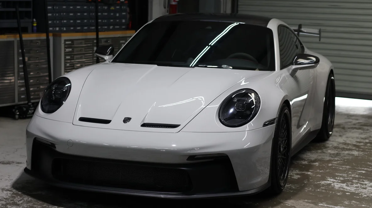 A white Porsche 911 sports car parked in a garage with tool cabinets in the background, showcasing its sleek design.