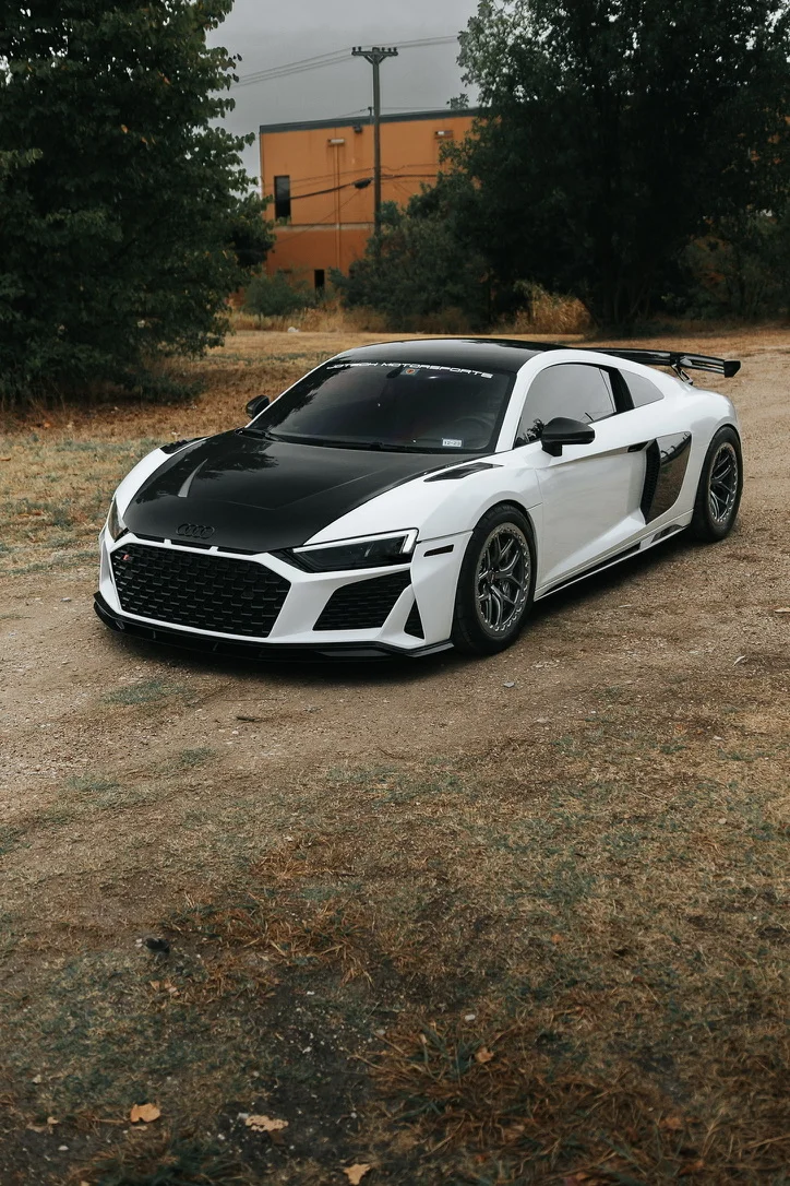 White Audi R8 with black carbon fiber hood, aftermarket wheels, and a large rear wing parked on a dirt road.