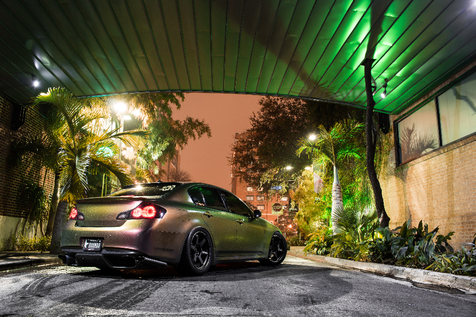 An iridescent Infiniti G37 sedan with aftermarket wheels parked at night under green lighting in an urban setting.