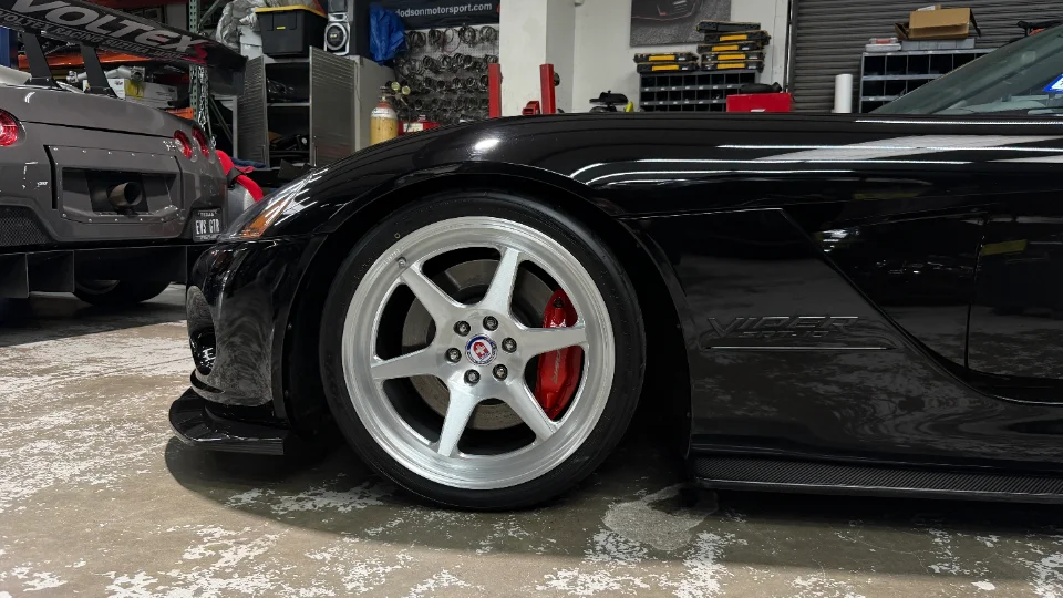 A black Dodge Viper with silver HRE wheels and red brake calipers parked in a performance shop, with a grey Nissan GT-R in the background.