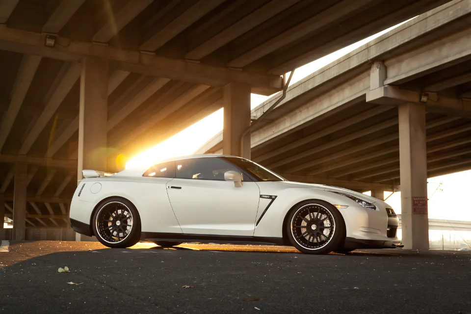 A white Nissan GT-R with aftermarket black wheels parked under an overpass during sunset, with a bright lens flare.