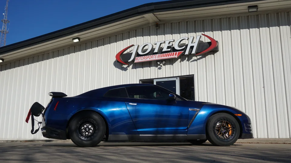 A blue Nissan GT-R drag car with a parachute parked outside the Jotech Motorsports facility on a sunny day.