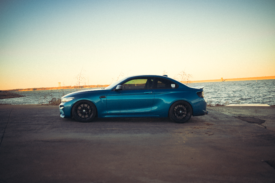 A blue BMW M2 coupe with black wheels parked by a lake at sunset.
