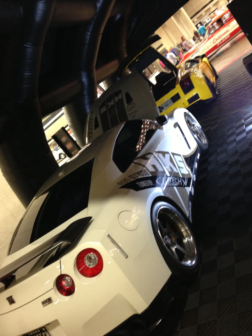 A white Nissan GT-R and a yellow Lamborghini Gallardo on display at an indoor car show, featuring custom wheels and HKS branding.