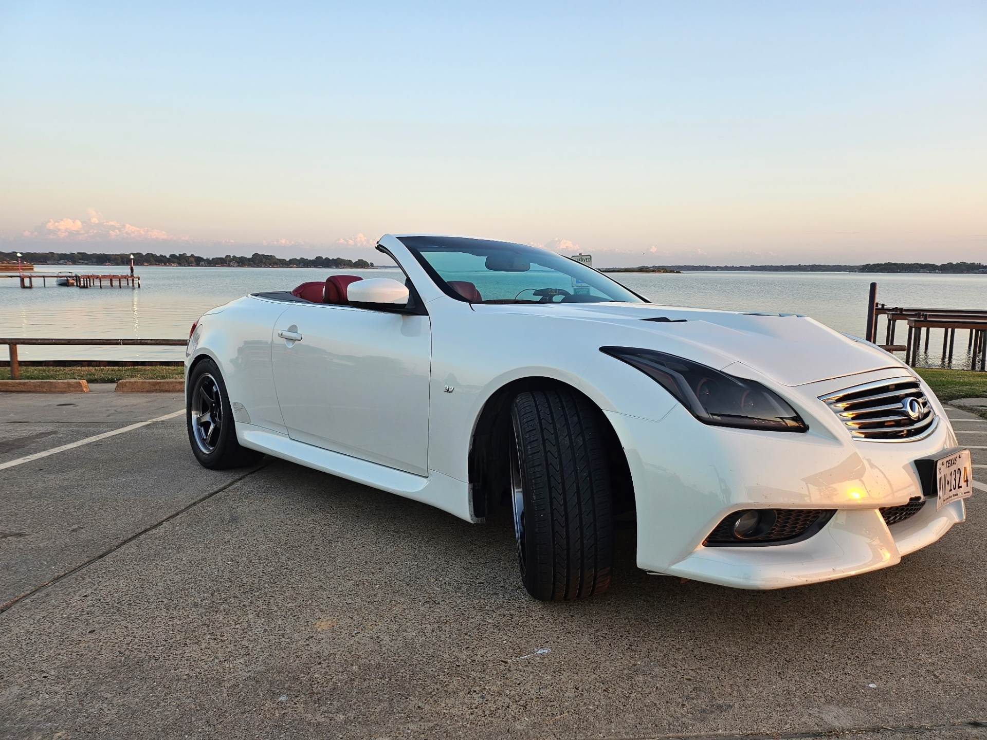A white Infiniti G37 convertible with aftermarket wheels parked by a lake during sunset.
