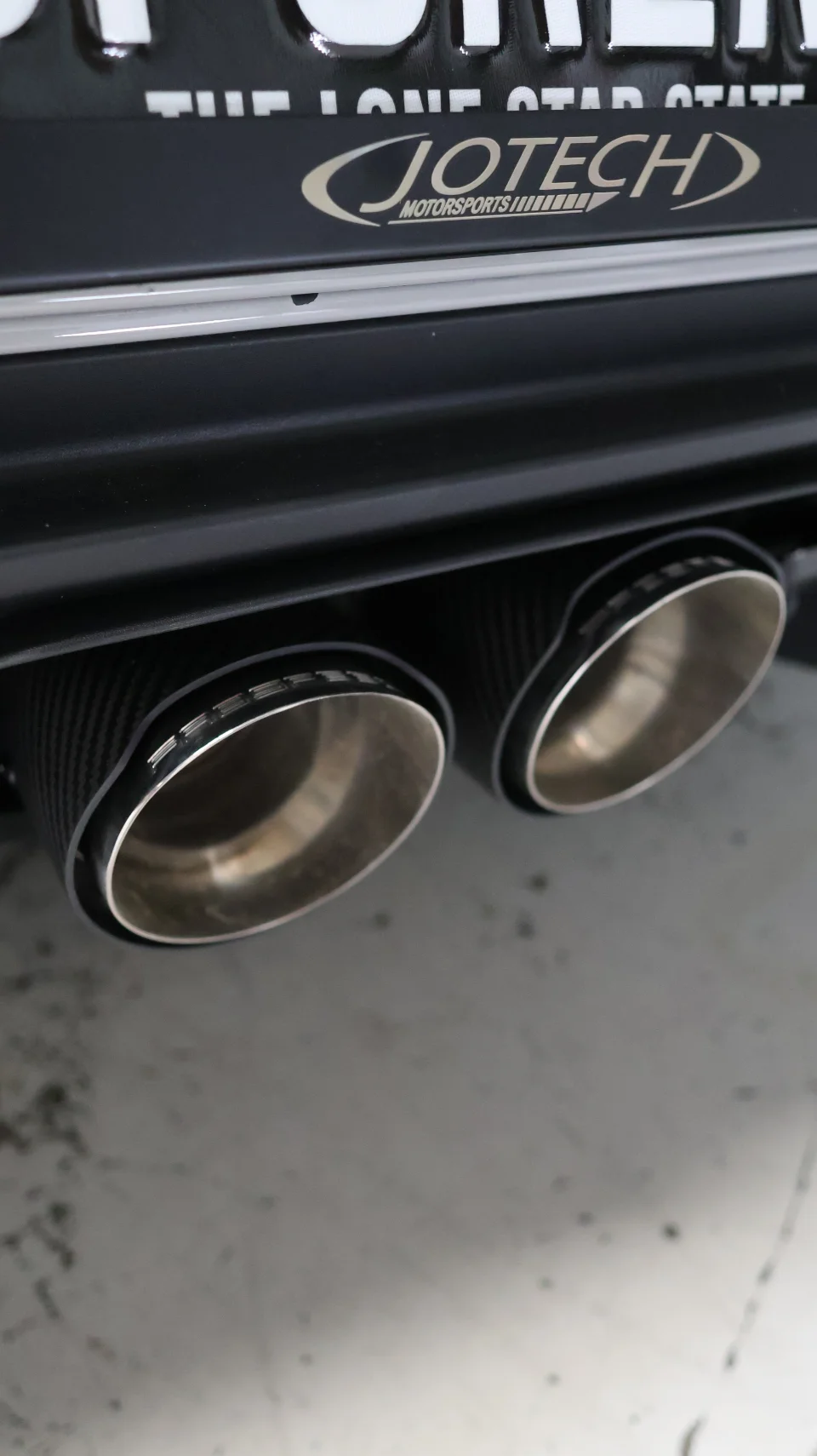 Close-up of carbon fiber exhaust tips on a performance car with a Jotech Motorsports license plate frame.
