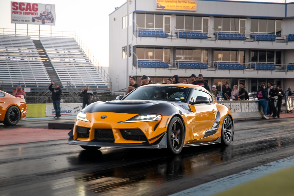Orange and black Toyota Supra on a drag strip with grandstands and a timing tower in the background