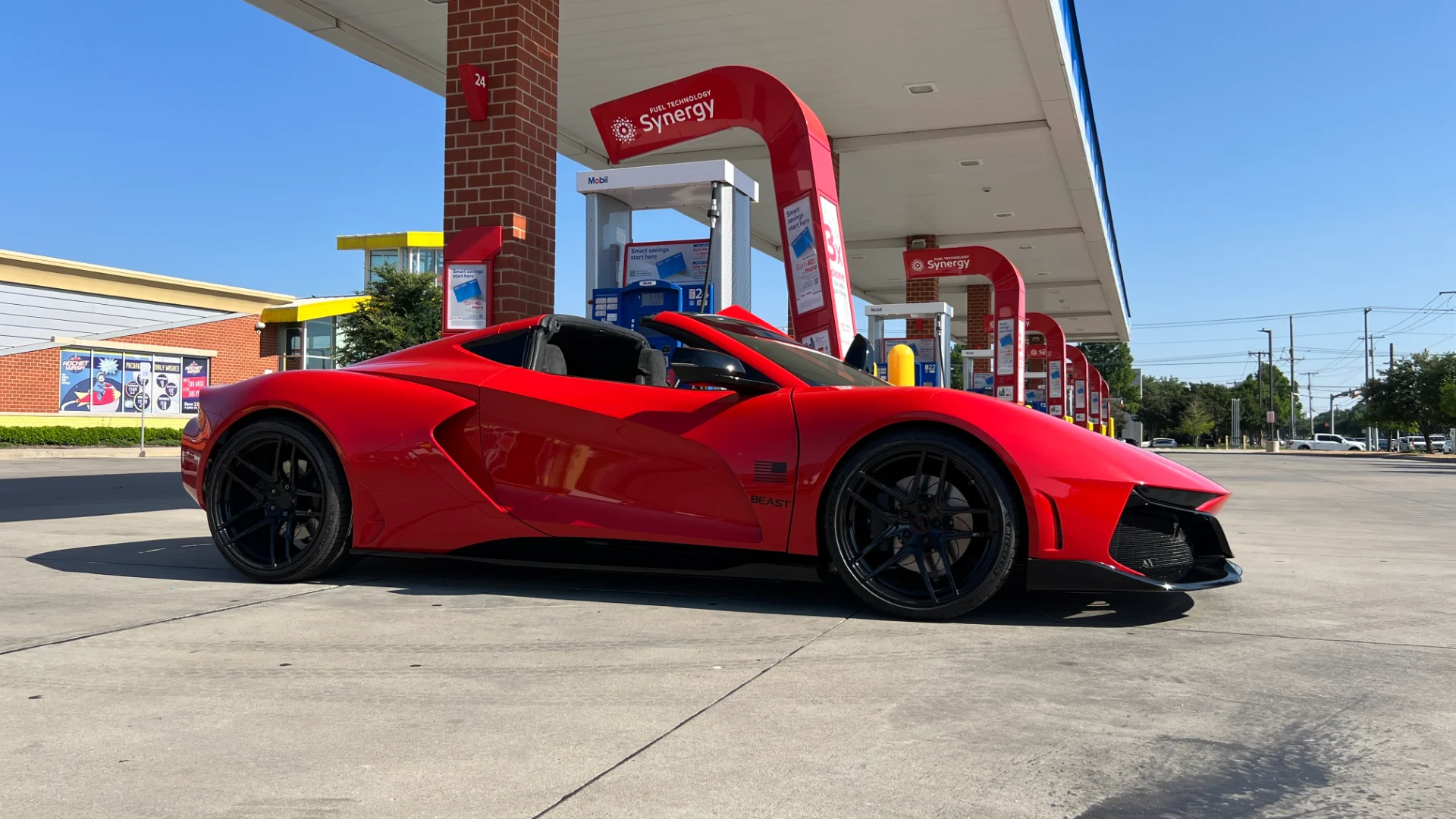 A striking red Valarra Beast custom sports car with black wheels parked at an Exxon gas station on a clear, sunny day.