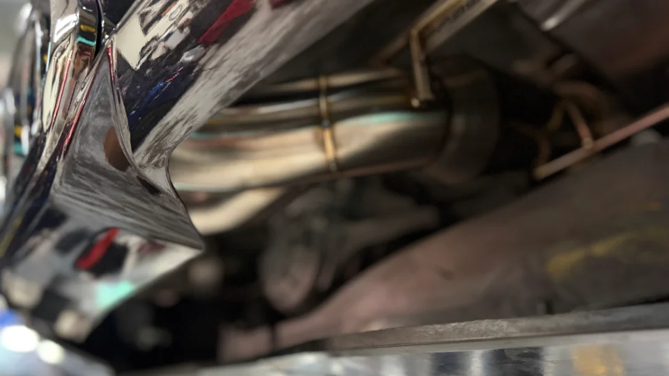 Close-up of a performance stainless steel exhaust system with visible welds on the underside of a car in a shop.