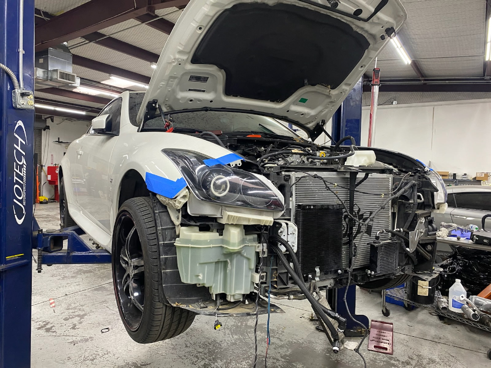 White Infiniti G37 coupe on a lift in a Jotech Motorsports shop with the front bumper removed for performance upgrades.