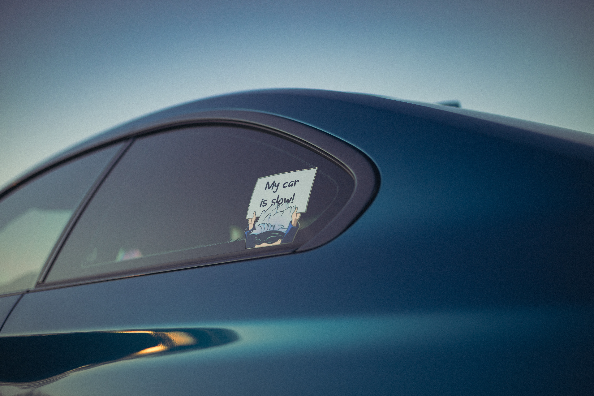 A close-up of the rear quarter panel and window of a teal car with a 'My car is slow!' sticker.