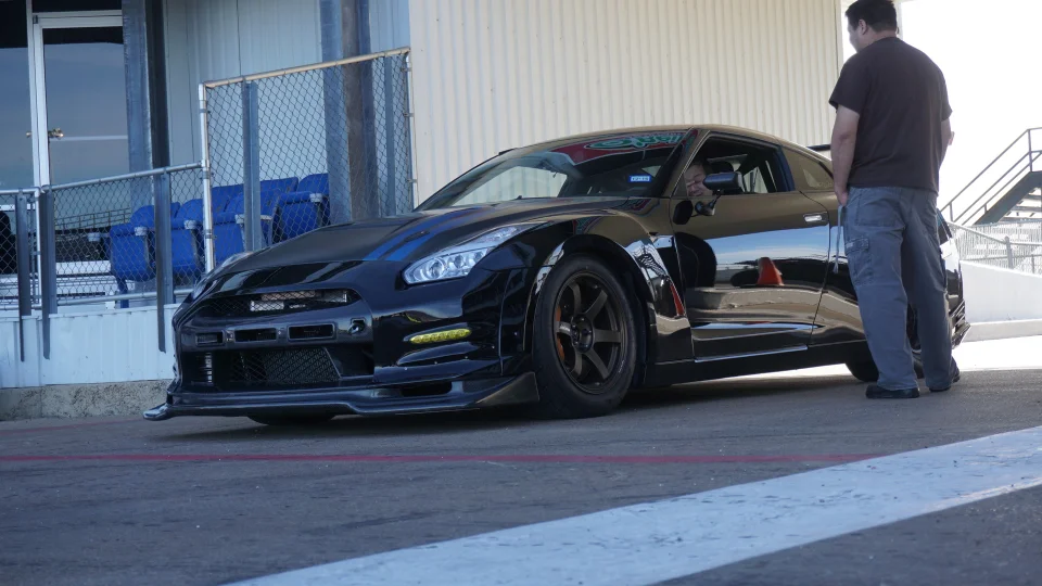 Black Nissan GT-R with aftermarket wheels and aero kit parked at a track, with a person standing next to it.