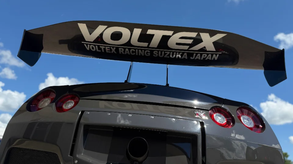 Rear view of a grey Nissan GT-R R35 with a large carbon fiber Voltex racing wing against a blue sky.