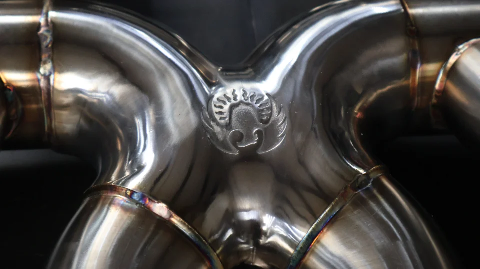 Close-up of a polished stainless steel exhaust x-pipe with visible welds and a custom logo.