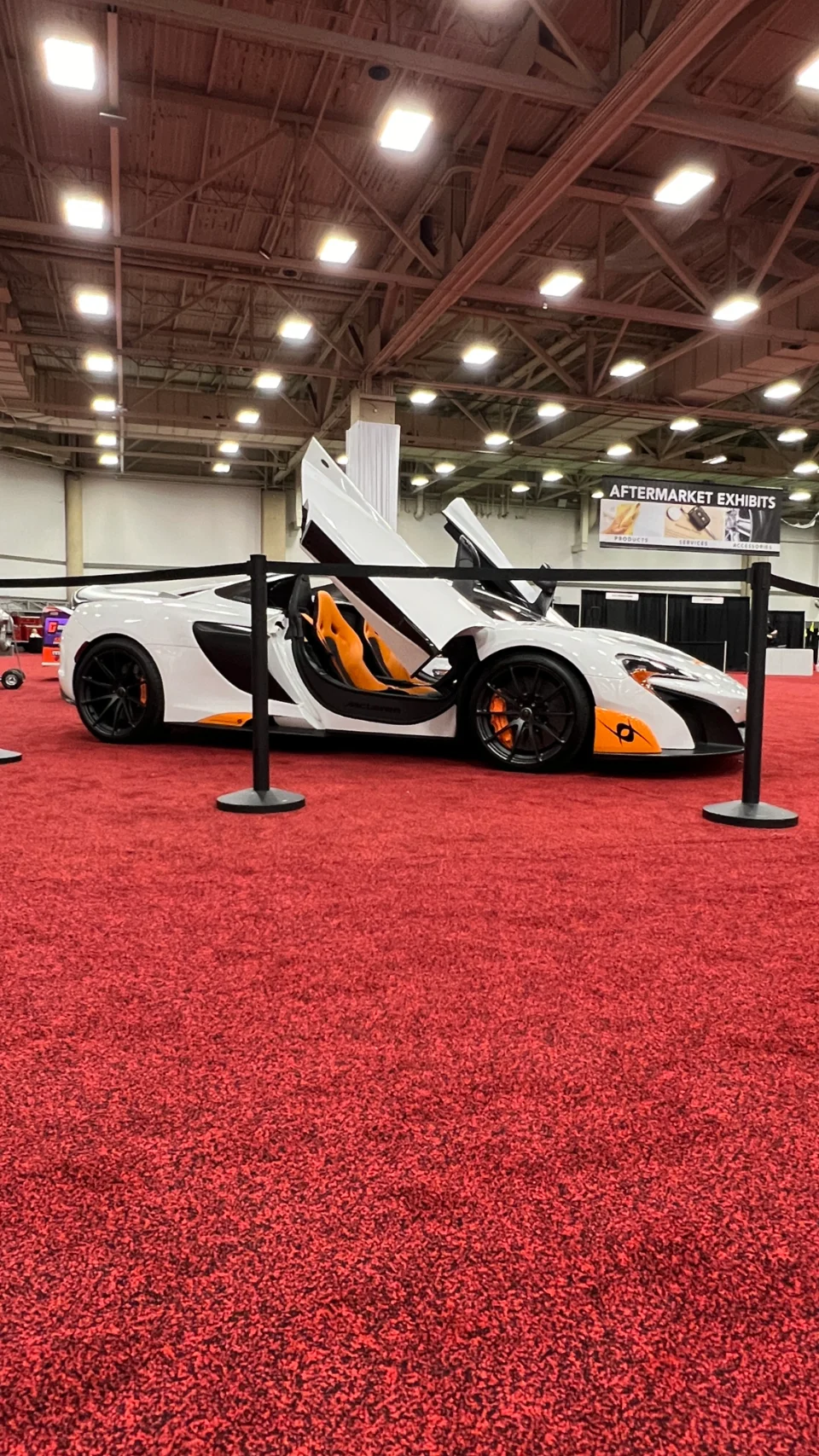 A white McLaren supercar with orange interior and open doors on display at a car show on a red carpet.
