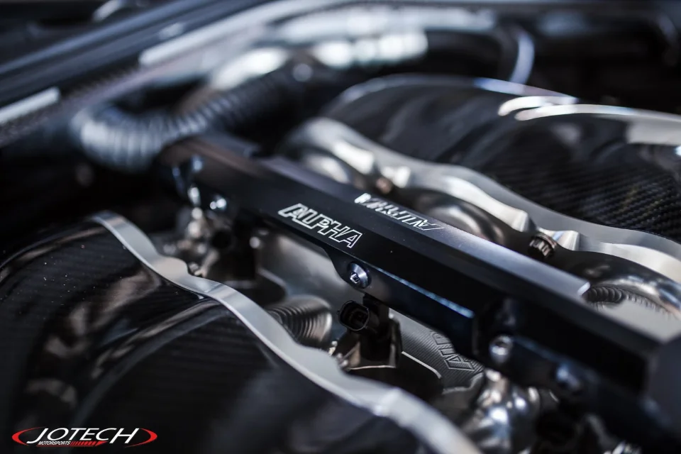 Close-up of Alpha Performance fuel rails in a car's engine bay, featuring carbon fiber components and Jotech Motorsports branding.
