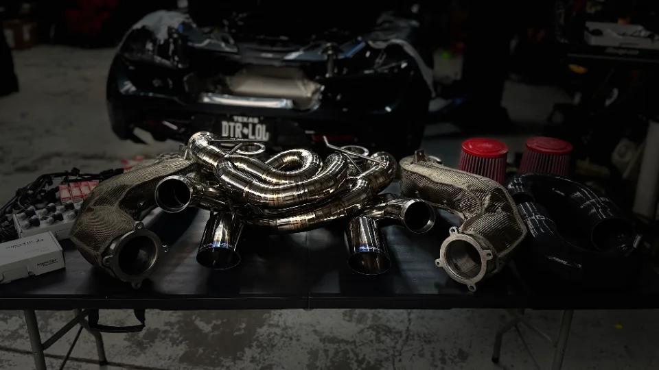 Performance exhaust headers, air filters, and other tuning parts laid out on a table in a shop with a McLaren in the background.
