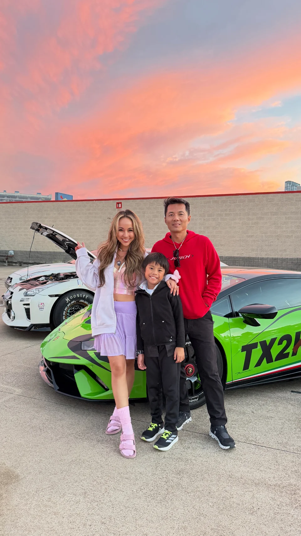A family poses with a green Lamborghini Huracan race car with TX2K livery at a track event during sunset.