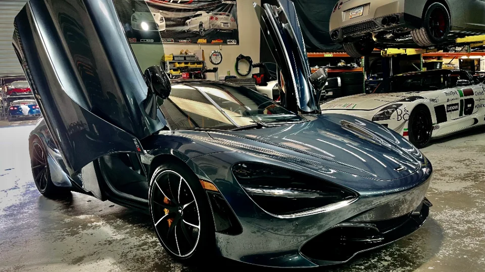 A dark grey McLaren 720S with its butterfly doors open, parked on a shop floor with other performance cars and equipment.