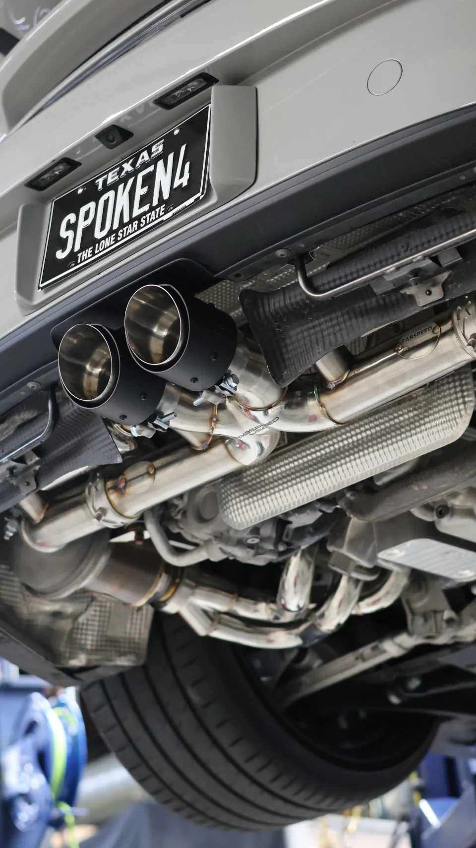 Detailed view of a Fabspeed exhaust system with black tips installed on a grey Porsche, seen from underneath while on a lift.