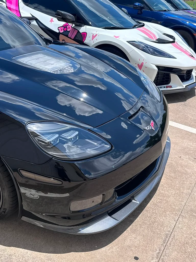 A black Corvette C6 with a carbon fiber front splitter parked next to a white Corvette C8 with a pink anime-style wrap at a car meet.