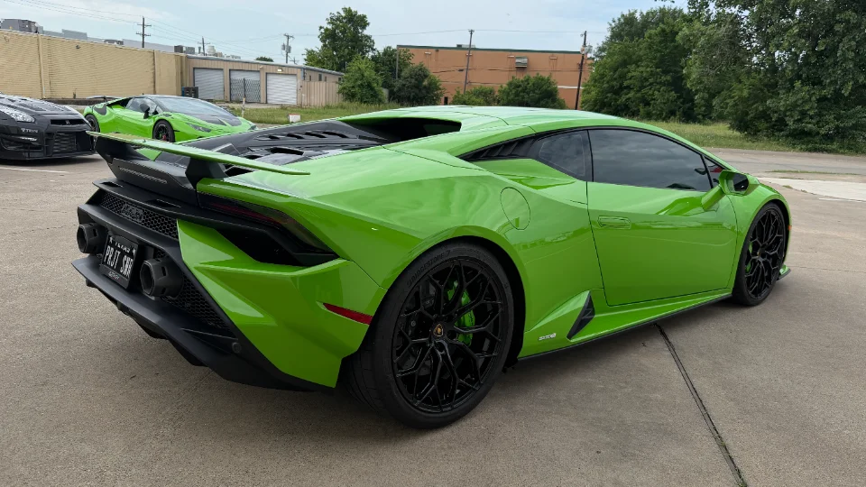 A vibrant green Lamborghini Huracan with an aftermarket rear wing parked on asphalt, with a black Nissan GT-R visible in the background.