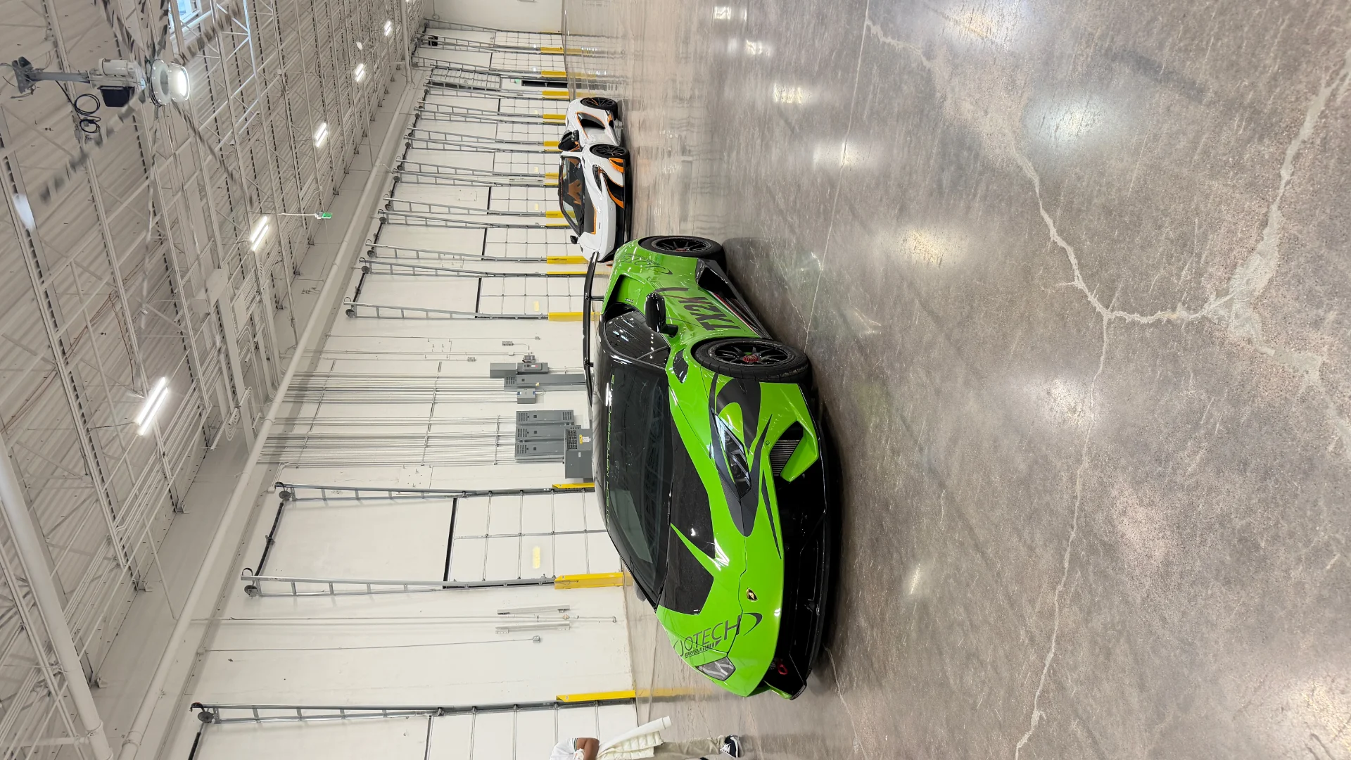 A vibrant green Lamborghini Huracan and a white McLaren supercar parked on a polished concrete floor in a large, modern facility.