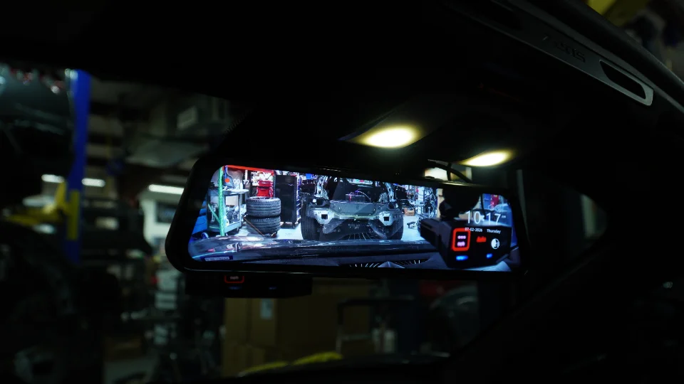 View from inside a car showing a digital rearview mirror displaying a disassembled car in a performance shop.