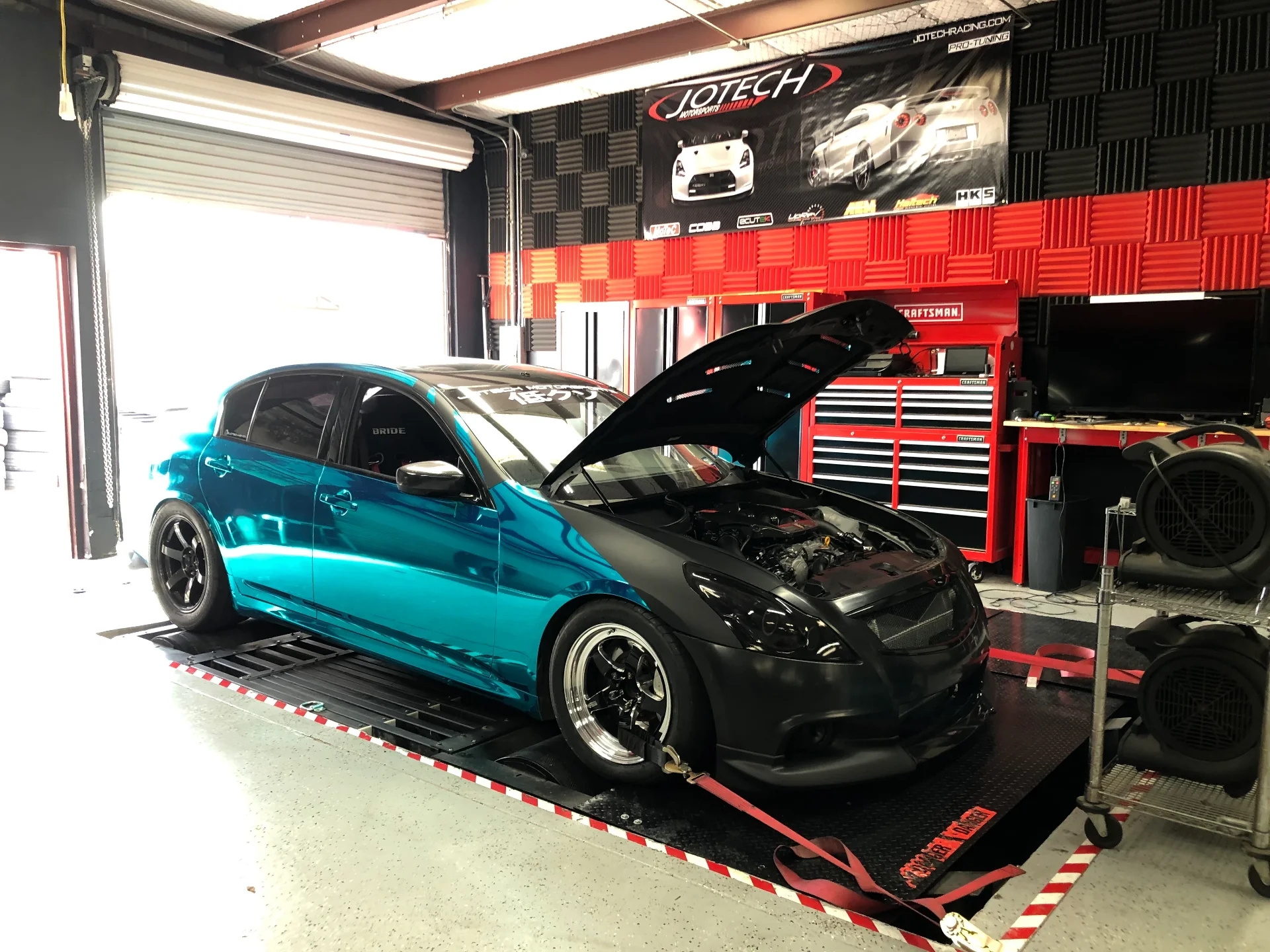 A teal and black Infiniti G37 hatchback strapped to a dynamometer at Jotech Motorsports for performance tuning.