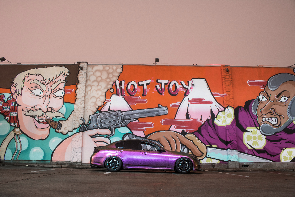 A custom purple wrapped Infiniti G37 sedan parked at night in front of a vibrant graffiti mural.