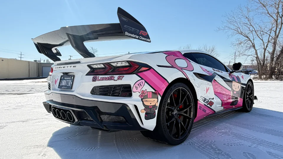 A white Chevrolet Corvette C8 with a custom pink anime-style wrap and a large aftermarket carbon fiber rear wing parked in a snowy lot.