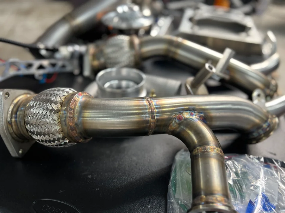 Close-up of custom fabricated stainless steel exhaust downpipes with visible welds and a flex pipe, laid out on a surface.