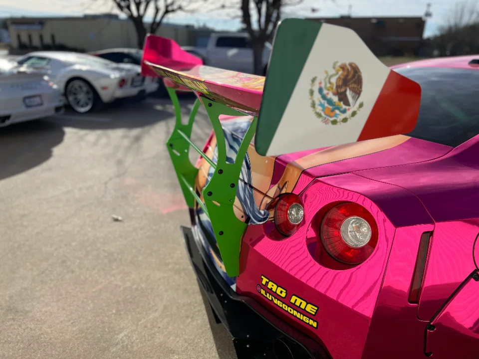 Rear view of a pink chrome wrapped Nissan GT-R with a large green custom rear wing featuring a Mexican flag design and anime graphics.