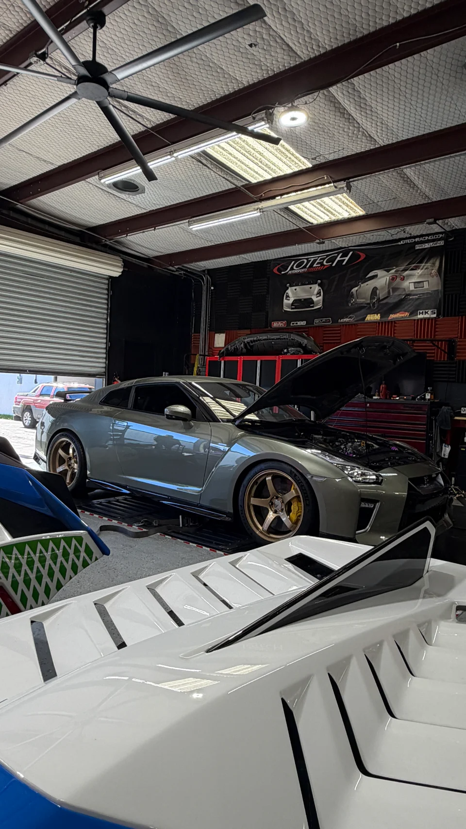 A grey Nissan GT-R with its hood open sits on a dyno at Jotech Motorsports, ready for tuning in the performance shop.