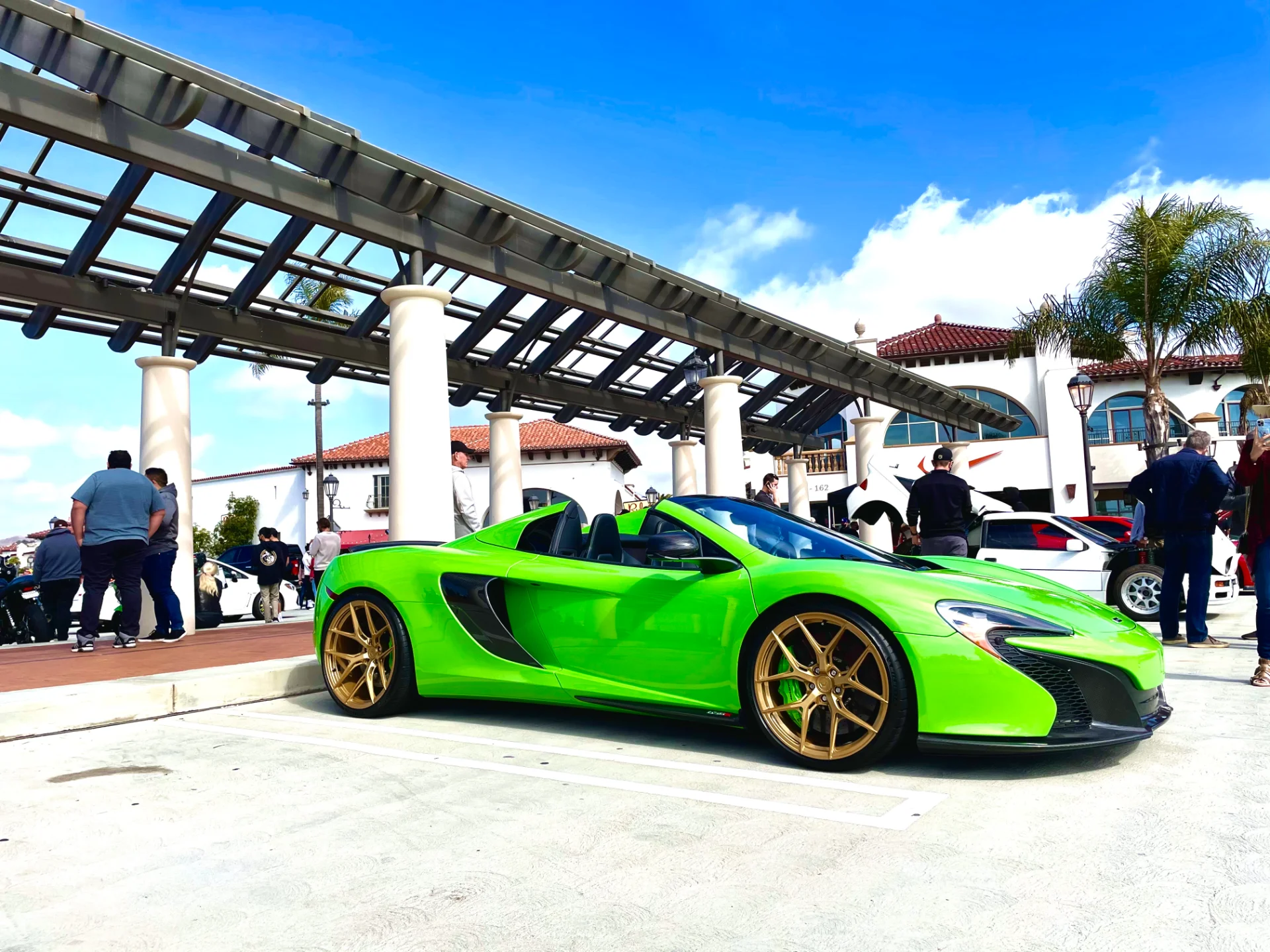 A bright green McLaren 650S Spider with gold wheels parked at an outdoor car meet under a blue sky.
