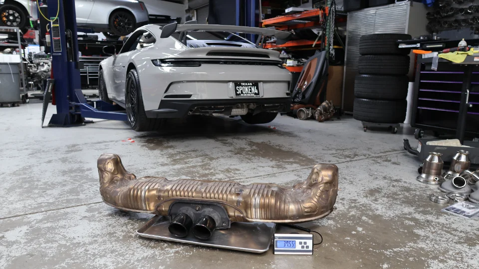 A grey Porsche 911 GT3 on a lift in a shop, with its stock exhaust system being weighed on a scale in the foreground.