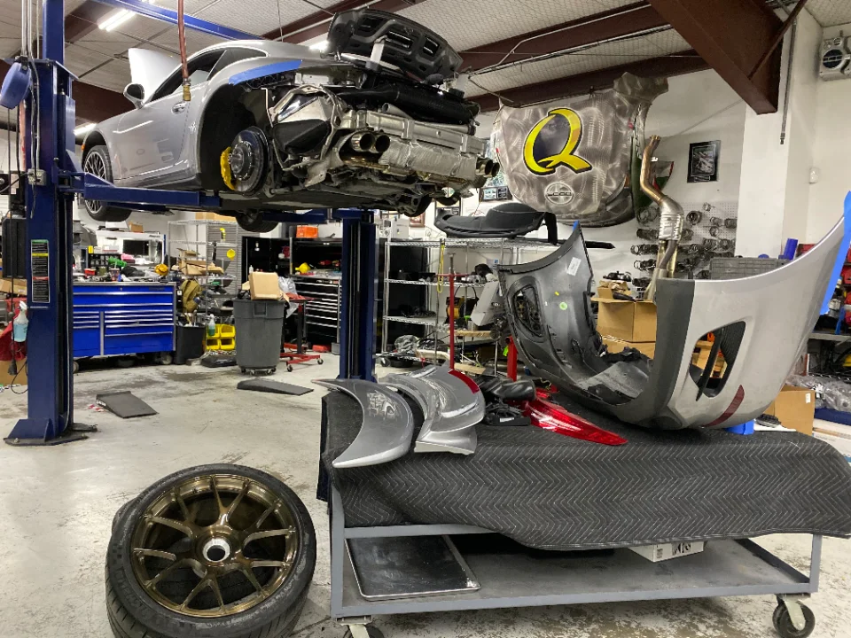 A silver Porsche 911 on a lift in a Jotech Motorsports shop, undergoing disassembly with its rear bumper and wheel removed.