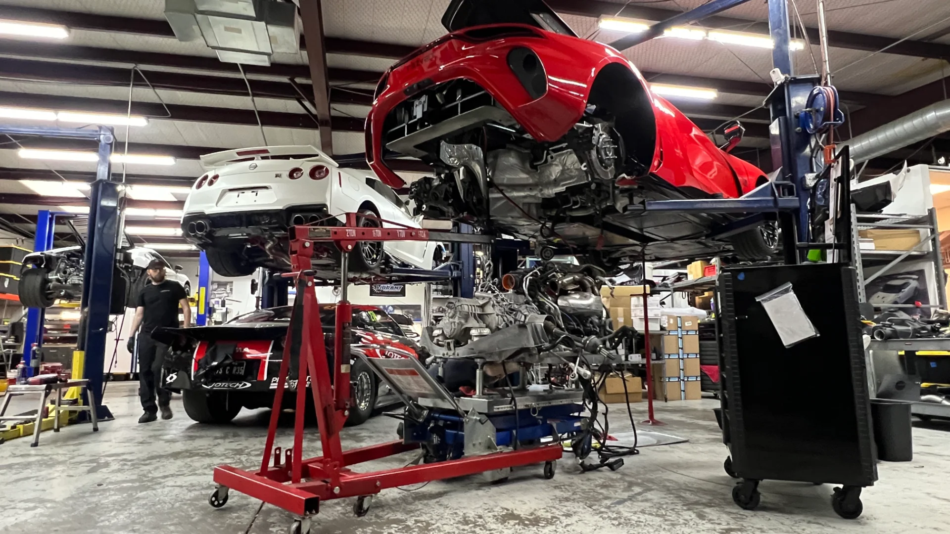 A red sports car and a white Nissan GT-R on lifts in an auto shop with an engine removed and a mechanic working.