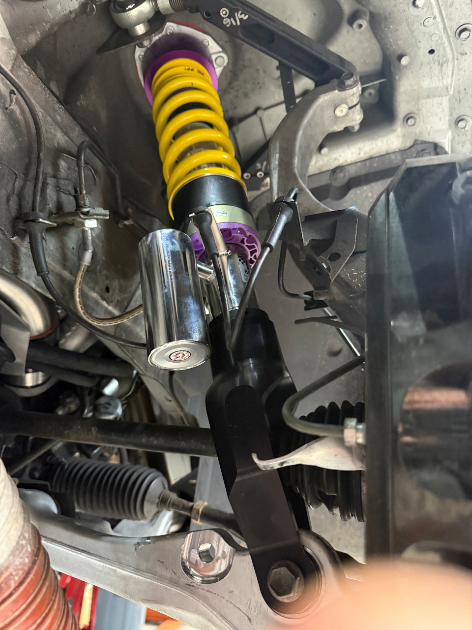A close-up view of a KW coilover suspension system with a yellow spring and purple components installed on a car's chassis.