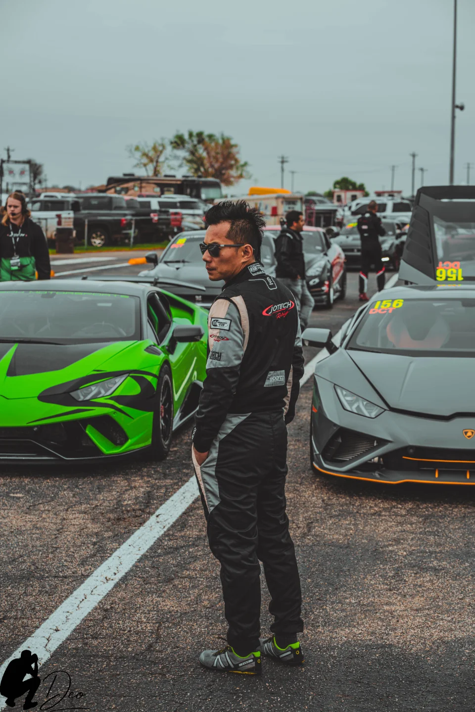 A man in a racing suit stands between a green Lamborghini and a gray Lamborghini at a drag strip.