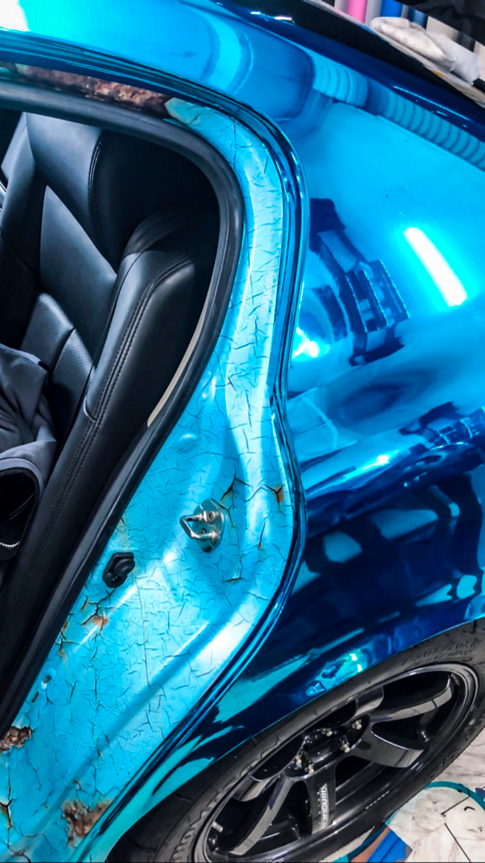 Close-up of a car's rear door jamb with a unique cracked blue chrome wrap, showing a black multi-spoke wheel and black leather interior.