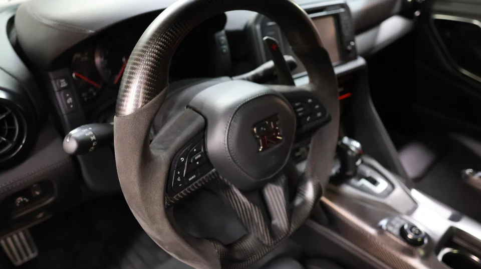 A close-up of a Nissan GT-R's interior, showing the carbon fiber and Alcantara steering wheel with paddle shifters.
